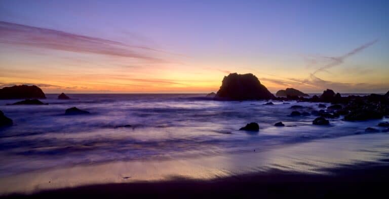 A purply sunet sky reflected in the pacific with black rocks